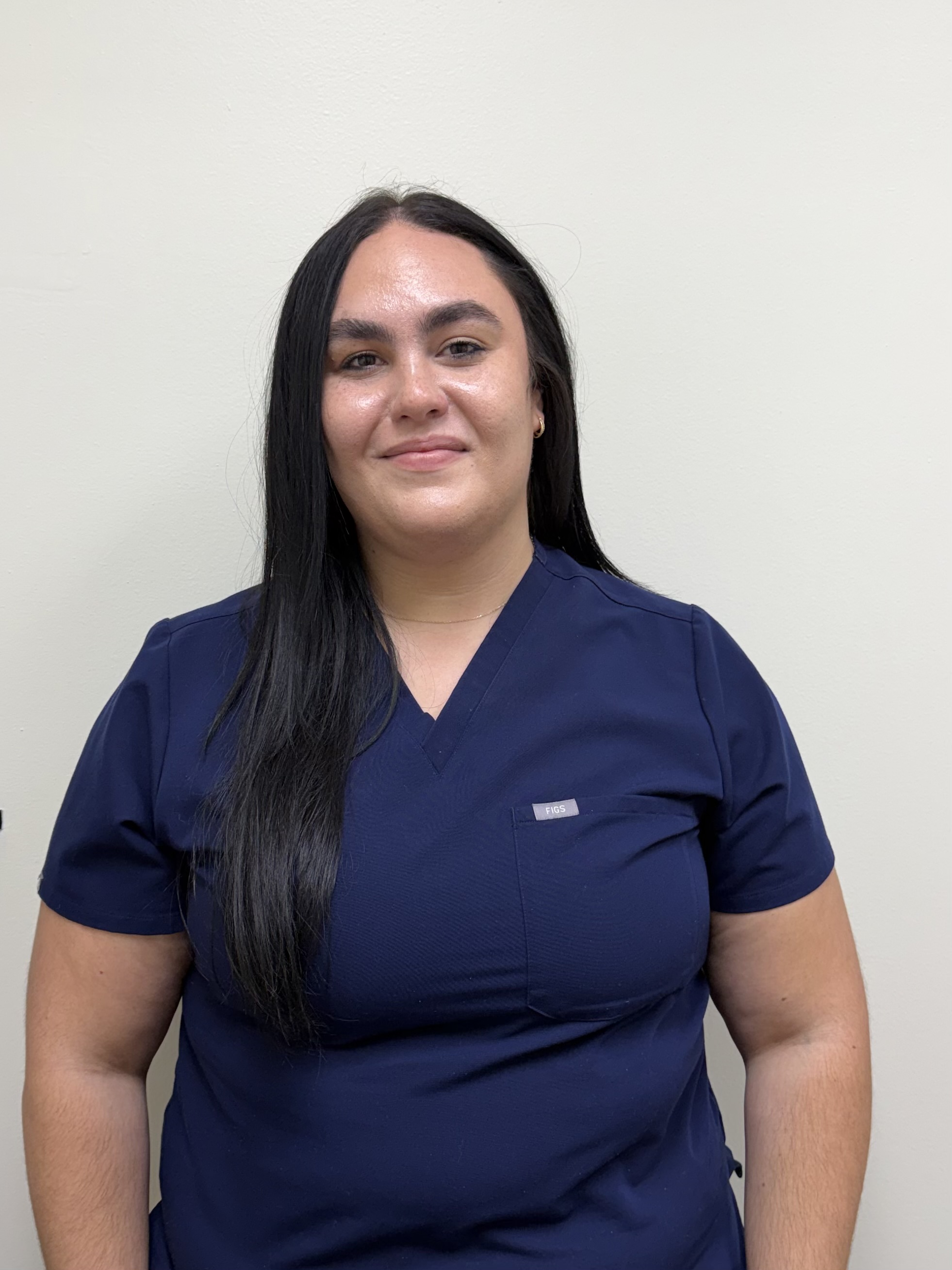 A woman with long dark hair wearing a blue scrub top stands against a plain background.