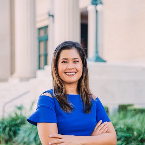 A woman standing in front of a building with columns, wearing a blue top and smiling at the camera.