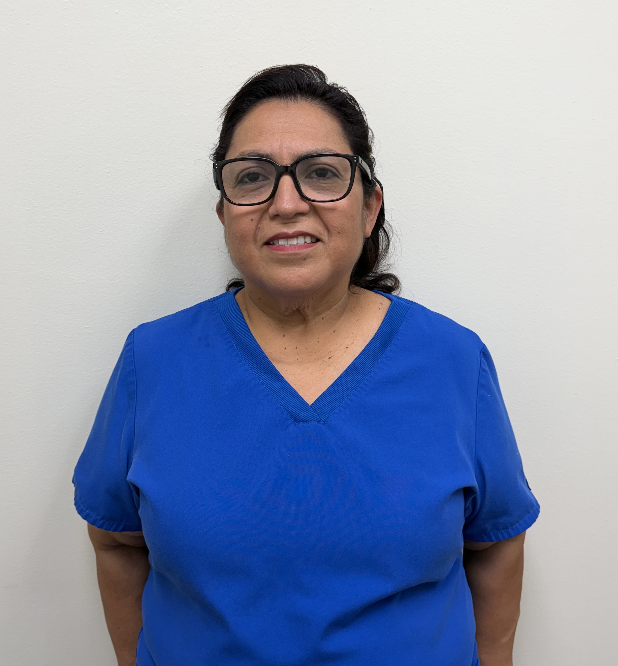 The image shows a woman wearing glasses, standing against a white wall, dressed in a blue scrub top and dark pants, with her arms crossed in front of her.