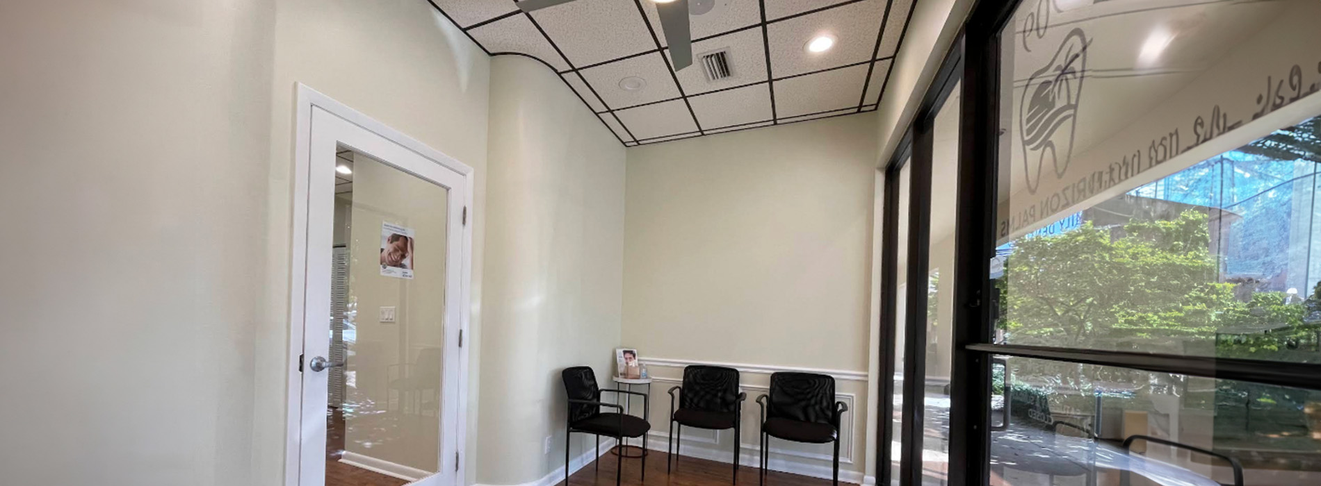 The image shows an interior space with a large window allowing natural light into the room, where there are chairs, a desk, and a wall-mounted TV.