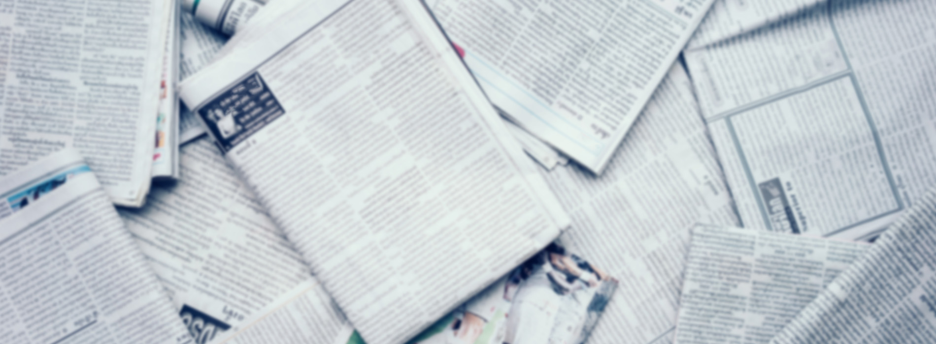 The image shows a collection of newspaper pages scattered on a surface with a focus on a central cluster of papers, creating an abstract pattern against a neutral background.