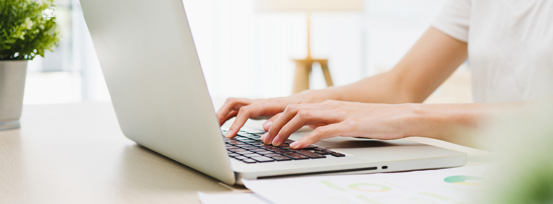 The image shows a person using a laptop computer on a table with a blurred background featuring another person working on a different computer.