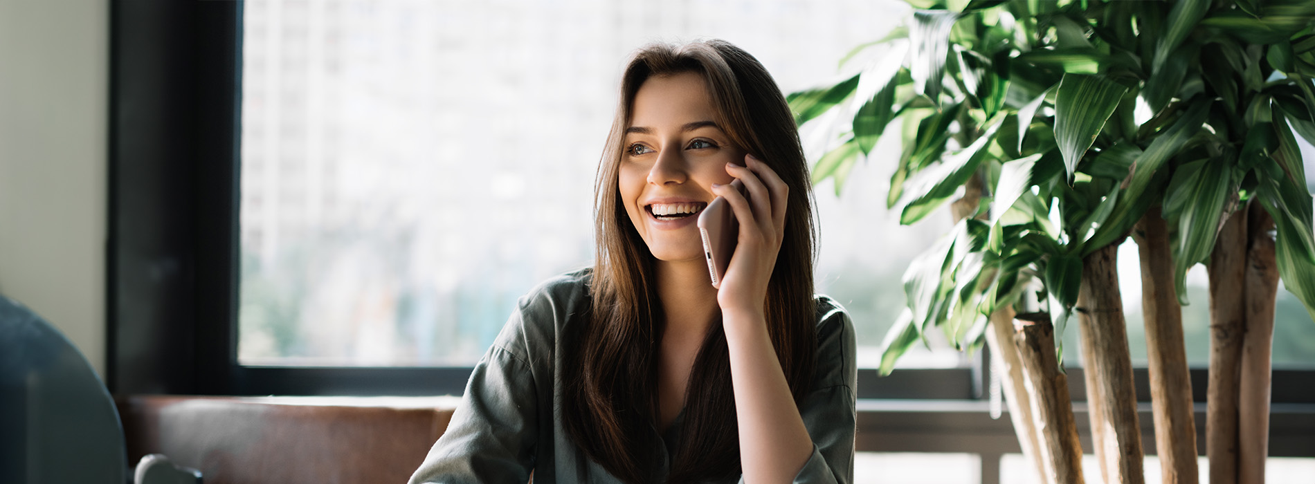 The image depicts a woman sitting at a table with a cell phone, smiling and looking towards the camera, set against an indoor background of a potted plant and a wall-mounted light fixture.