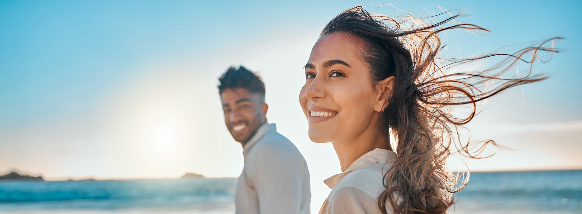 The image shows a man and woman standing on a beach with their arms around each other, smiling and looking at the camera.