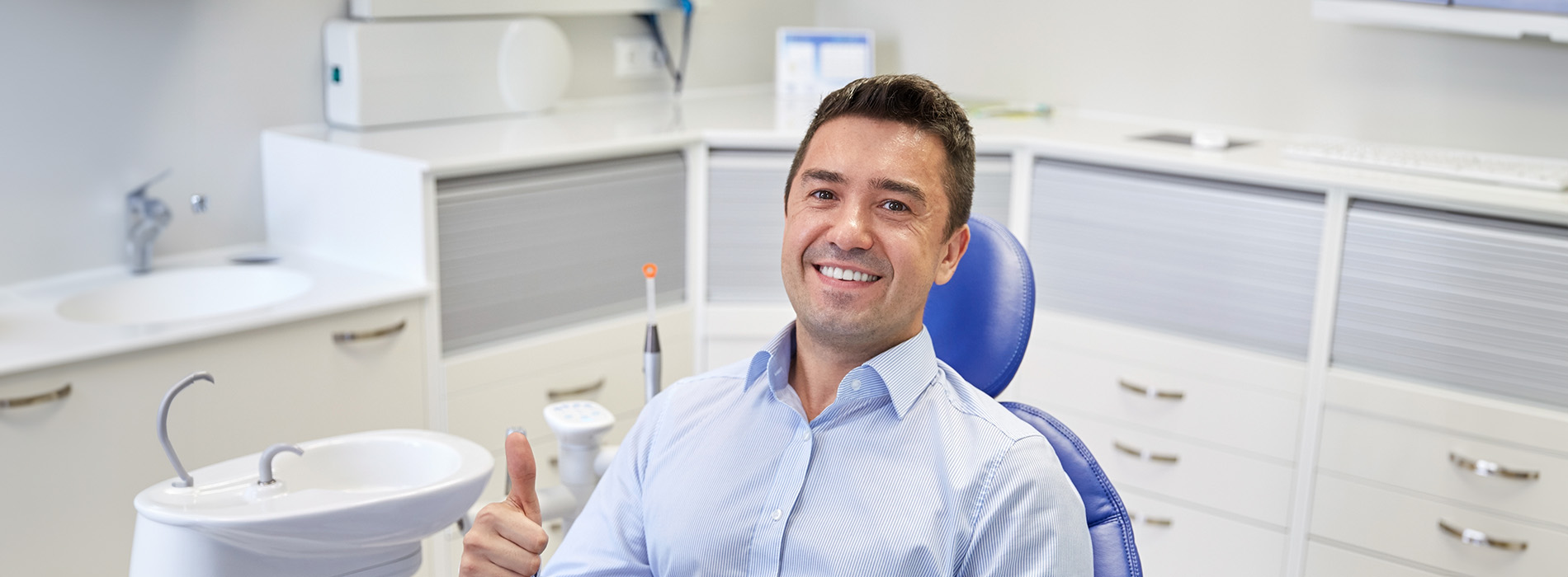 The image shows a man sitting at a dental chair with a thumbs-up gesture. He is wearing a blue shirt, and behind him is a well-equipped dental office setting with cabinets, equipment, and a sink.