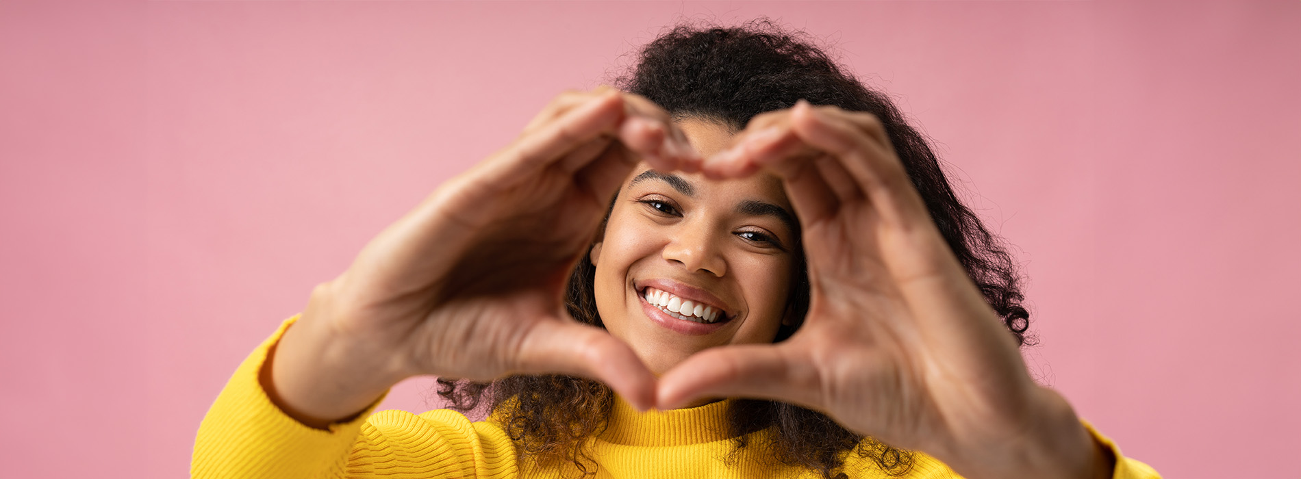 The image shows a person making a heart sign with their hands against a pink background.