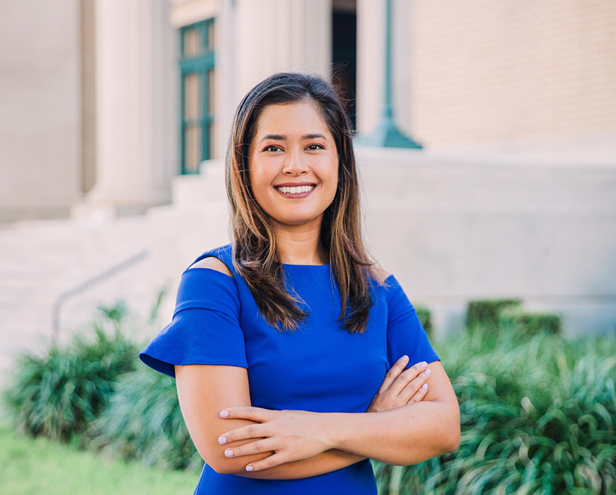 The image shows a woman standing outdoors in front of a building with columns, wearing a blue dress and smiling at the camera.