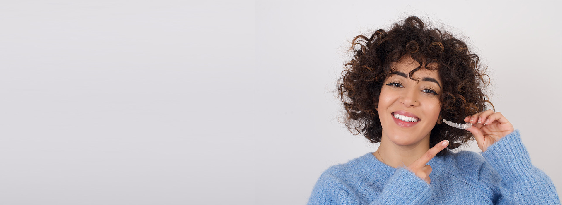 The image features a woman with curly hair smiling at the camera while holding a toothbrush to her mouth. She has dark skin and is wearing a blue sweater, and there s a watermark on the right side of the photo.