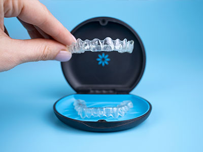 The image shows a person s hand holding an open dental impression tray with a set of transparent aligners inside, placed on a blue surface against a white background.