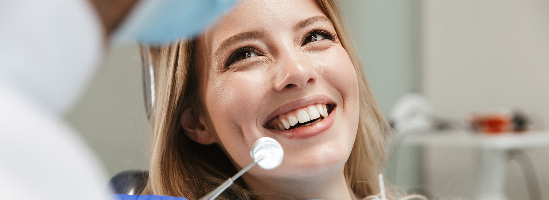 A young woman with a bright smile sits in a dental chair, looking directly at the camera, while a dentist examines her teeth.