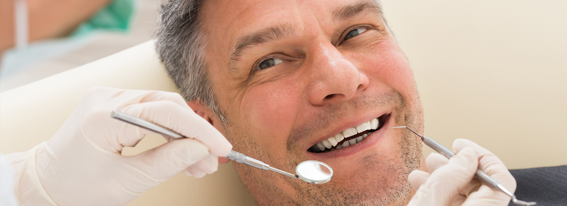 The image shows a man with a beard sitting in a dental chair, receiving dental treatment from a dentist who is cleaning his teeth with a dental tool.