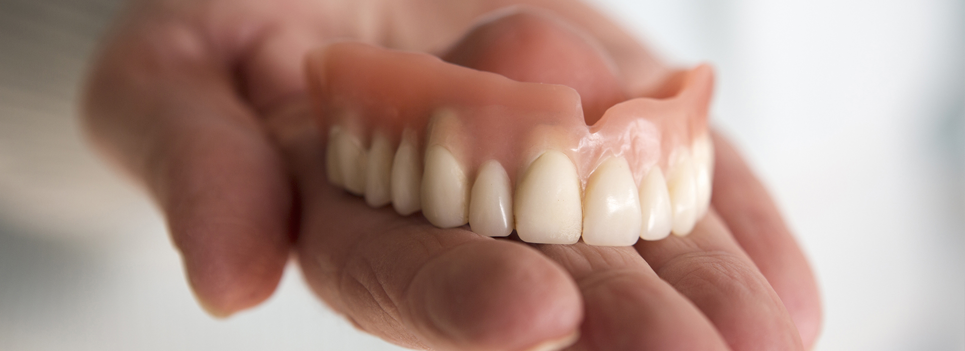 A human hand holding up a set of dentures with visible teeth, against a white background.