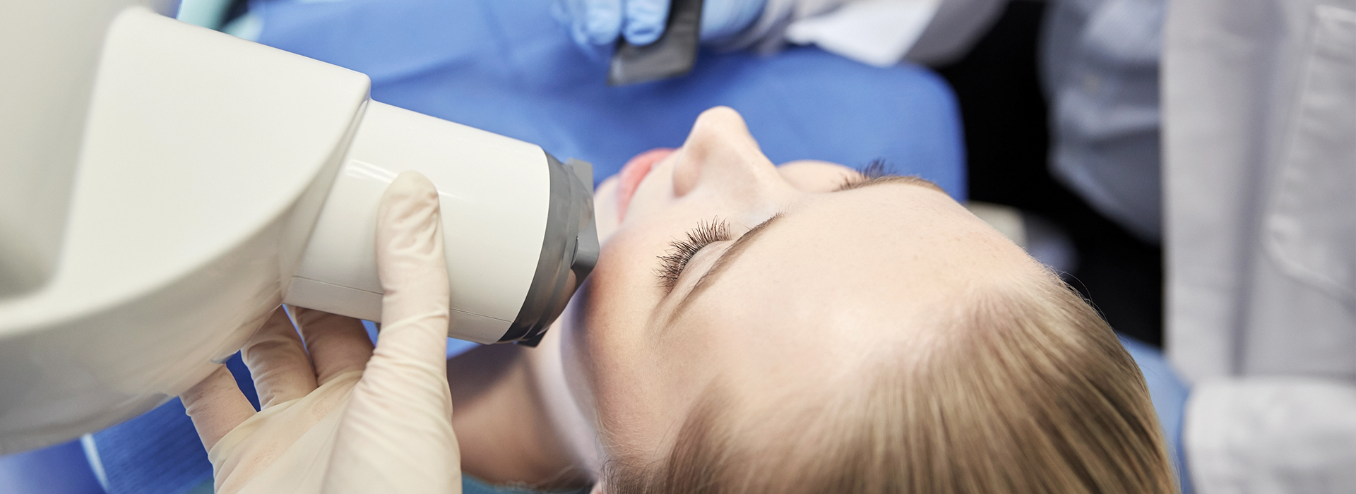 The image shows a person having their hair cut under a magnifying device, with a medical setting background featuring blue curtains and medical equipment.