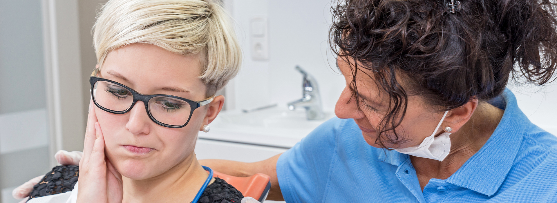 The image shows a woman wearing glasses and holding her face with one hand while another person, possibly a dental professional, is examining her teeth with a mirror.