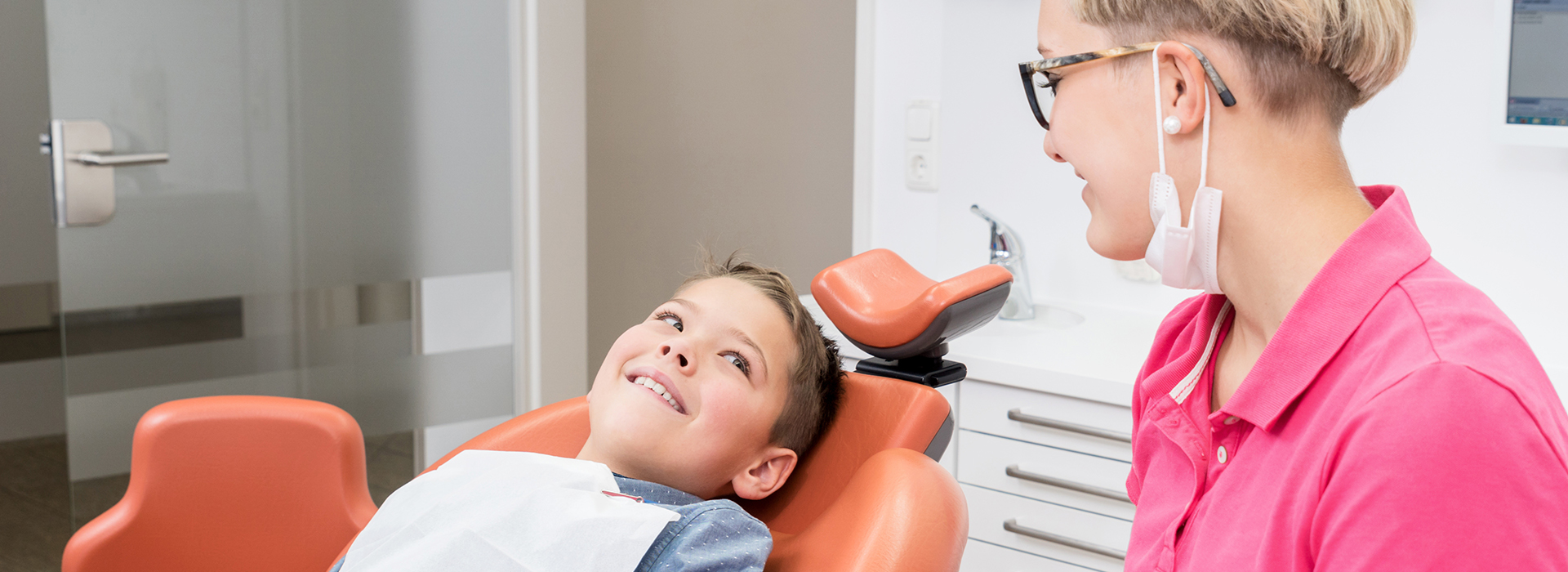 The image shows a young boy sitting in a dental chair with his eyes closed while a woman, presumably a dentist, attends to him. They are both in an office setting.