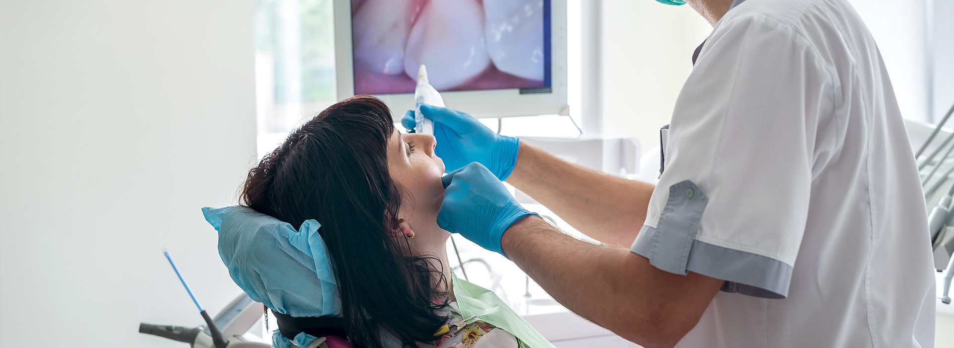 The image shows a dental professional adjusting a patient s teeth with their hands while wearing gloves, set against a blurred background featuring medical equipment and a person sitting down.