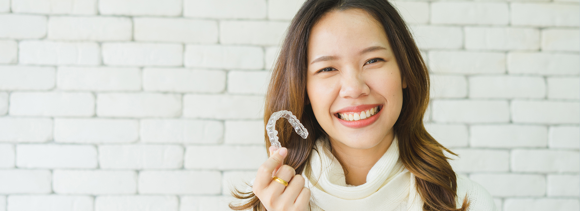 A young woman with a bright smile holds up a toothbrush against her face, standing indoors with a brick wall behind her.