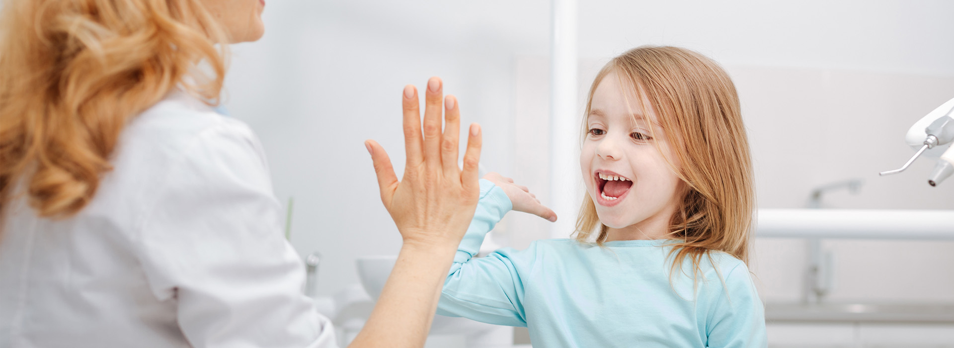 The image shows a young girl with blonde hair clapping her hands in front of an adult woman who appears to be giving her high-fives. They are both indoors, likely in a bathroom setting, as suggested by the presence of a sink and mirror in the background.