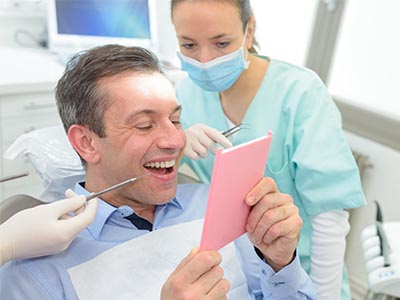 The image shows a man sitting in a dental chair with a large smile, looking at his reflection in a mirror while holding up a pink card with text on it, surrounded by dental equipment and a dental professional standing behind him.