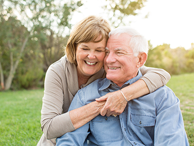 This is a color photograph featuring an elderly couple   a man and a woman   embracing each other outdoors during daylight hours, with both individuals smiling and looking at the camera.
