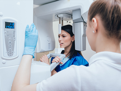 In this image, there are two individuals, presumably healthcare professionals, standing in front of a CT scan machine. The person on the right is wearing a blue protective gown and appears to be operating the scanner, while the one on the left, dressed in a white lab coat, is looking at the screen with interest or concern.