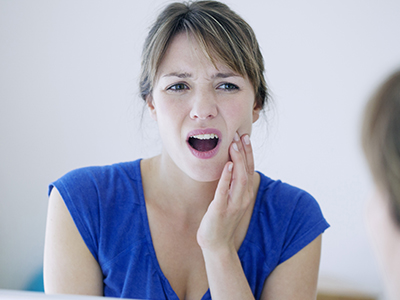The image shows a woman with her mouth open, displaying teeth, while looking at a mirror with an expression of surprise or concern.