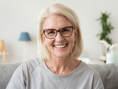 The image shows a woman with short blonde hair smiling at the camera. She has glasses on her face and appears to be indoors, possibly in a living room setting.