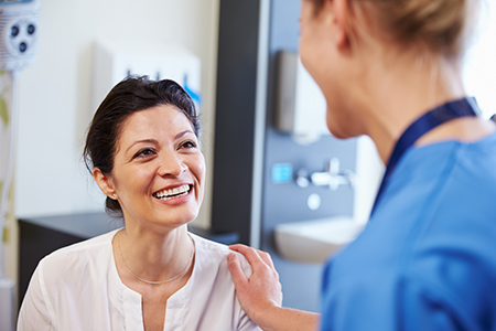 The image shows two individuals, one woman with a smile looking at another person who appears to be a healthcare professional, possibly a nurse, in an indoor setting that suggests a medical environment.