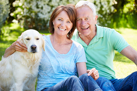 The image shows an elderly couple sitting outdoors with a dog on their laps, posing for a photograph.