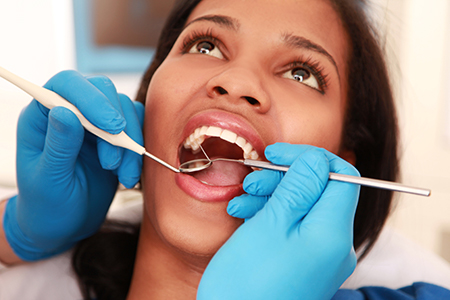 In the image, there is a woman sitting in a dental chair with her mouth open, receiving dental treatment from a dentist wearing blue gloves and using dental instruments.