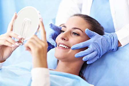 A woman sits under a magnifying mirror, with a medical professional adjusting her glasses, both in a clinical setting.