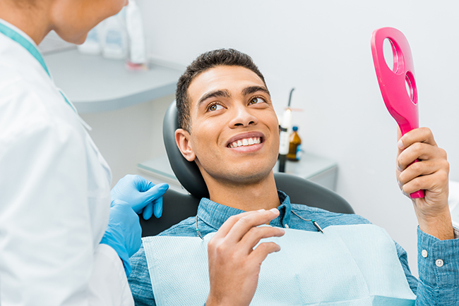 A man sitting in a dental chair with a dentist s mask on his face, smiling at the camera while holding a pink object in his hand.
