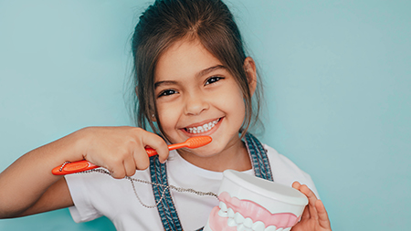 A young girl brushing her teeth with a red toothbrush, smiling at the camera while holding a toothpaste tube.