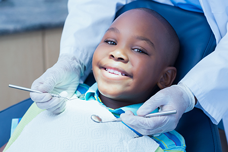 The image shows a young boy sitting in a dental chair with a broad smile, receiving dental care from a professional wearing gloves and a face mask.