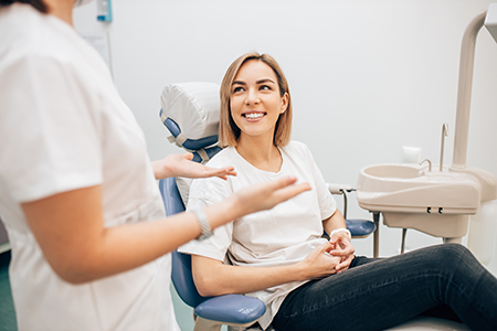 The image shows a woman sitting in a dental chair with a smile on her face, being attended to by a dental professional in a clinical setting.
