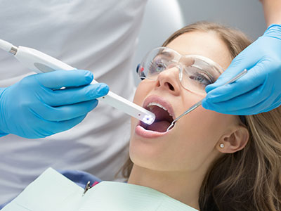 A woman receiving dental treatment with a dental drill being operated by a dentist wearing blue gloves and using an electric device, while she holds her mouth open wide for the procedure.