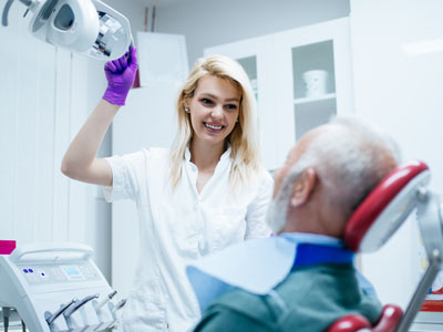 A woman in a white lab coat stands next to an older man with a medical mask on his face, both are inside a medical room filled with equipment.