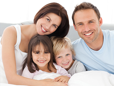 A family poses together on a couch with smiles, featuring an adult couple with three children.