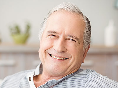 This is a portrait-style photograph of an older man with gray hair, smiling and looking directly at the camera. He appears relaxed and content, wearing a dark shirt and seated indoors.