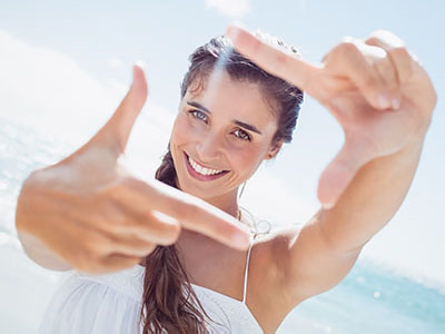 The image shows a woman taking a selfie at the beach with her hand held up against the camera lens, smiling and looking directly into the camera.