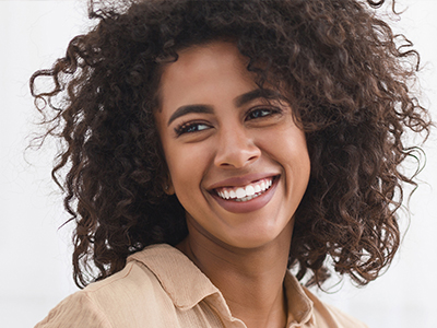 The image shows a person with curly hair smiling at the camera, wearing a light-colored top and standing against a plain background.